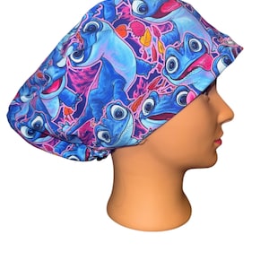 May include: A blue and pink patterned scrub cap featuring a cartoon character design. The cap is made of a soft fabric and is designed to fit comfortably. The design includes blue characters with pink accents and autumn leaves.