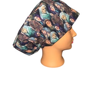 May include: A patterned surgical scrub cap featuring a repeating design of various faces in shades of brown, green, and purple. The cap is worn on a mannequin head, showcasing its shape and fit. The fabric appears to be cotton or a similar material.