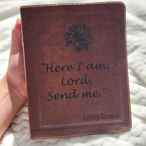 May include: A brown leather-bound journal with the quote "Here I am, Lord, Send me." and the name "Emily Grace" engraved on the cover. A floral design is above the quote. The journal is held against a white, textured background.