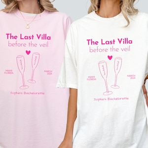 May include: Two white t-shirts with pink text and graphics. The text reads "The Last Villa before the veil". The graphic is two champagne flutes with the text "Sophia's Bachelorette" below. The t-shirts are on two women.