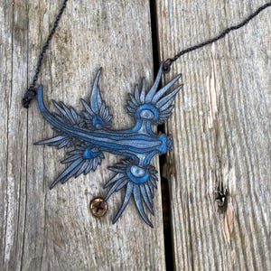 May include: A blue and gray pendant necklace featuring a stylized sea creature design. The pendant is suspended from a dark chain. The necklace is displayed on a weathered wooden surface.