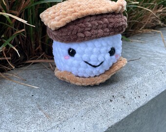 Könnte beinhalten: Ein handgefertigtes gehäkeltes S'more-Plüschtier. Das Plüschtier hat ein weißes Marshmallow mit einem lächelnden Gesicht, eine dunkelbraune Schokoladenschicht und hellbraune Graham Cracker. Das Spielzeug liegt auf einer Betonoberfläche mit grünem Gras im Hintergrund.