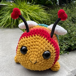 Puede incluir: Una abeja de crochet hecha a mano, con cuerpo amarillo, cabeza roja y rayas arcoíris. Tiene ojos grandes, negros y brillantes, una boca sonriente y antenas rojas en forma de corazón. Alas blancas en forma de nube están unidas a la espalda de la abeja.