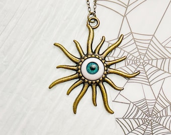May include: A gold-toned necklace with a sunburst pendant featuring a blue eye in the center. The pendant is surrounded by a white circle with a beaded edge.