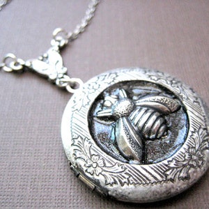 Puede incluir: Un collar con un medallón de plata con un diseño de abeja. El medallón tiene un patrón floral detallado alrededor del borde y un fondo gris oscuro.