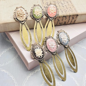 May include: Six antique-style metal bookmarks with oval cameo designs featuring a rose. The cameos are in various colors, including cream, green, pink, and blue. The bookmarks have a bronze finish and are arranged on a light-colored surface.