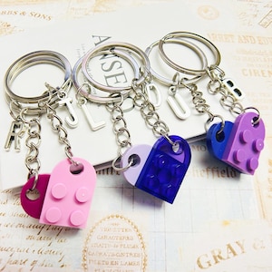 May include: Four silver keychains with letter charms and heart-shaped charms made of Lego bricks. The hearts are in shades of purple, pink, and blue.