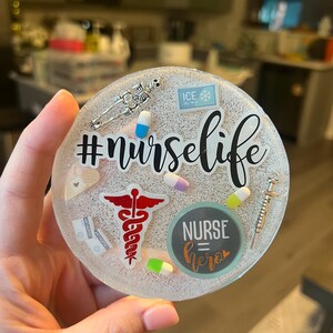 Könnte beinhalten: Ein runder, transparenter Harz-Untersetzer mit verschiedenen medizinischen Verzierungen, darunter ein Caduceus, Pillen, eine Spritze, ein Verband und ein Skelett. Der Text "#nurselife" und "NURSE=hero" ist ebenfalls auf dem Untersetzer enthalten.
