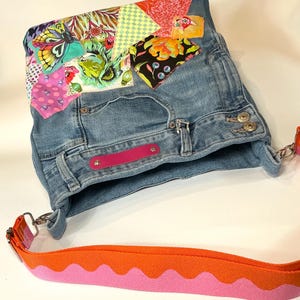 May include: A blue denim shoulder bag featuring a patchwork design of colorful fabric scraps with butterfly, lion, and floral patterns. The bag includes a bright pink rectangular accent and a wide orange and pink wavy-patterned strap.
