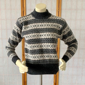 May include: A gray and cream-colored patterned sweater with a mock turtleneck. The sweater features a geometric design across the chest and sleeves. The cuffs and bottom hem are a darker gray color. The sweater is displayed on a mannequin.