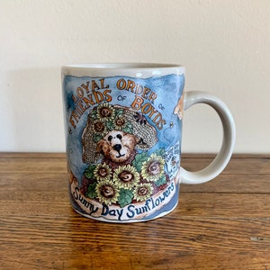 May include: A white ceramic mug with a blue and brown design featuring a teddy bear wearing a sunflower hat and surrounded by sunflowers. The mug has the text "Loyal Order of Boyd's Sunny Day Sunflowers" printed on it.