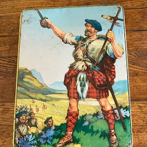 May include: A vintage tin box with a colorful illustration of a Scottish Highlander in a kilt, holding a sword and a staff. The Highlander is standing in a mountainous landscape with other figures in the background.
