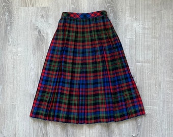 May include: A pleated, knee-length skirt with a classic plaid pattern in red, green, blue, and black. The skirt is made of a woven fabric and has a high waist. The skirt is displayed on a light gray wooden surface.