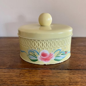 May include: A yellow metal tin with a floral design. The tin has a round shape and a lid with a knob. The floral design features a pink rose with green leaves and blue accents.
