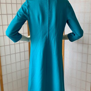 May include: A turquoise blue dress with a round neckline and three-quarter sleeves. The dress has a back zipper and a slightly flared skirt.