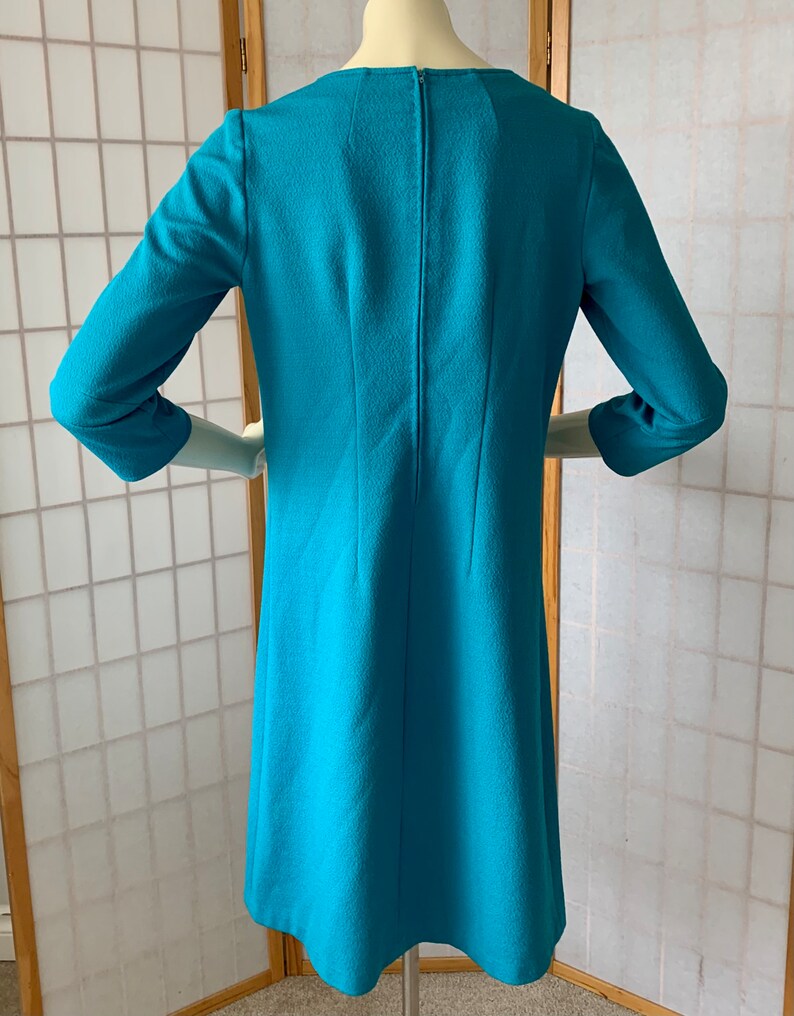 May include: A turquoise blue dress with a round neckline and three-quarter sleeves. The dress has a back zipper and a slightly flared skirt.