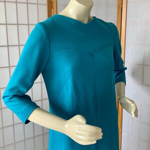 May include: A teal blue dress with a V-neck and three-quarter sleeves. The dress has a textured fabric and a fitted silhouette.