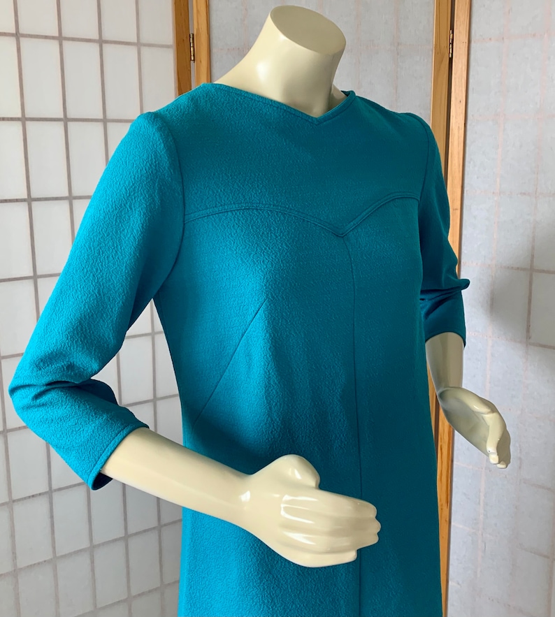 May include: A teal blue dress with a V-neck and three-quarter sleeves. The dress has a textured fabric and a fitted silhouette.