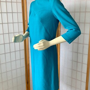May include: A turquoise blue dress with a V-neck and three-quarter sleeves. The dress has a fitted silhouette and a textured fabric.