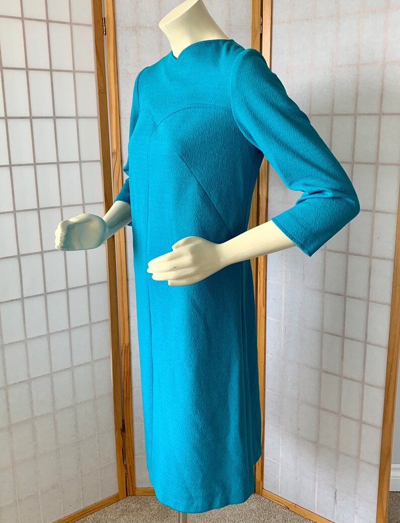 May include: A turquoise blue dress with a V-neck and three-quarter sleeves. The dress has a fitted silhouette and a textured fabric.