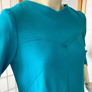 May include: A teal blue dress with a textured fabric and a V-neckline. The dress has a unique design with a seam that creates a heart shape in the front.