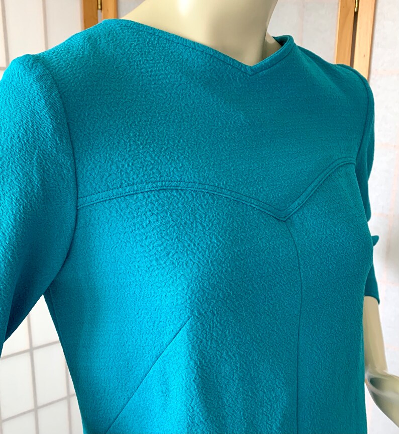 May include: A teal blue dress with a textured fabric and a V-neckline. The dress has a unique design with a seam that creates a heart shape in the front.