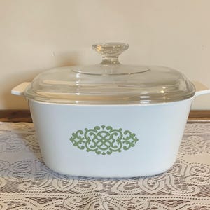 May include: A white casserole dish with a clear glass lid and handle. The dish features a green decorative design on the front. The lid has a clear glass knob. The dish is sitting on a white lace tablecloth.