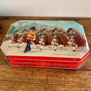 May include: A rectangular tin with a red plaid pattern on the sides and a lid featuring a colorful illustration of the Scots Guards marching in front of Edinburgh Castle. The tin is likely a collectible or souvenir item.
