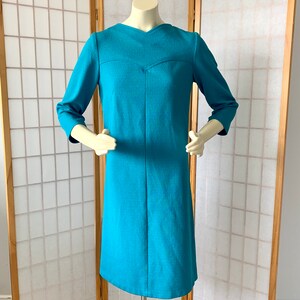 May include: A turquoise blue dress with a v-neck and three-quarter sleeves. The dress is made of a textured fabric and has a fitted silhouette.