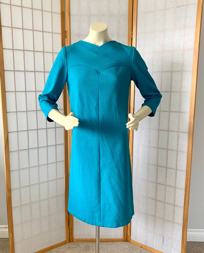 May include: A turquoise blue dress with a v-neck and three-quarter sleeves. The dress is made of a textured fabric and has a fitted silhouette.