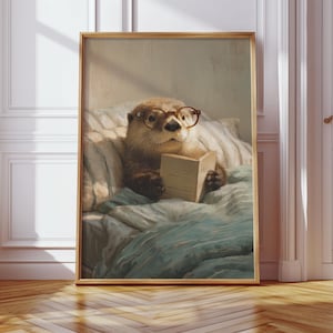 May include: A framed print featuring an otter wearing glasses and reading a book in bed. The otter is the focal point, set against a backdrop of soft bedding and a light-filled room. The frame is a warm, natural wood tone.