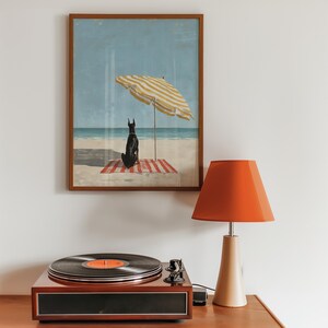 May include: A framed print depicts a black dog sitting on a red and white striped beach towel under a yellow and white striped umbrella. A vintage record player and an orange lamp sit on a wooden surface.