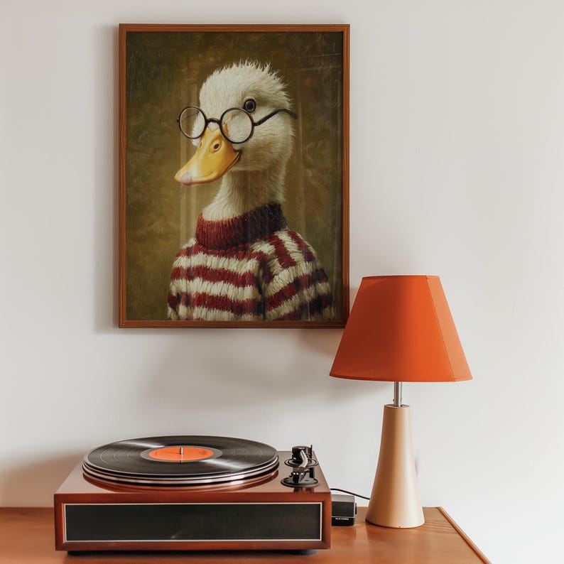 Op de afbeelding: Een ingelijst portret van een eend met een bril en een rood-witte gestreepte trui. Een platenspeler met een vinylplaat en een oranje lamp staan op een houten oppervlak.