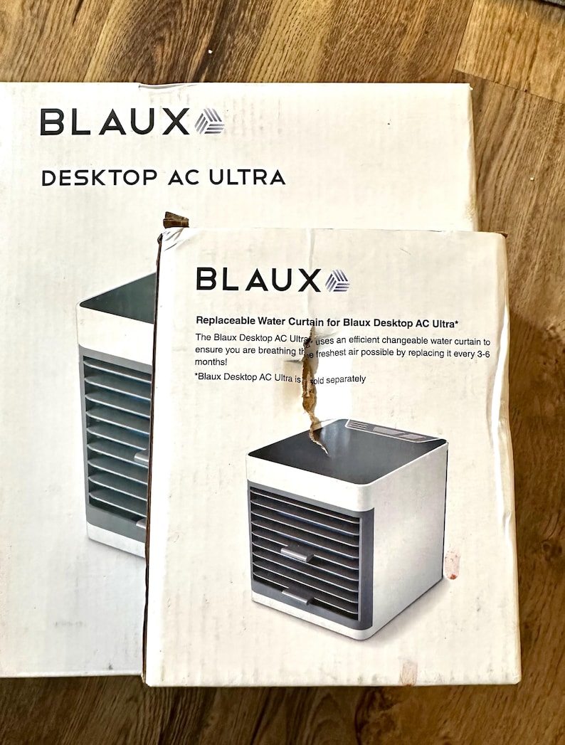 Puede incluir: Dos cajas blancas con el texto "BLAUX DESKTOP AC ULTRA" y "Replaceable Water Curtain for Blaux Desktop AC Ultra". Las cajas presentan una imagen de un peque&ntilde;o enfriador de aire blanco y gris.