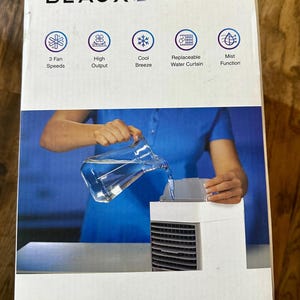 Puede incluir: Una caja de enfriador de aire port&aacute;til blanca y negra con la marca "BLAUX" en la parte superior. La caja presenta iconos que indican 3 velocidades de ventilador, alto rendimiento, brisa fresca, cortina de agua reemplazable y funci&oacute;n de niebla. Una persona vierte agua en el enfriador.