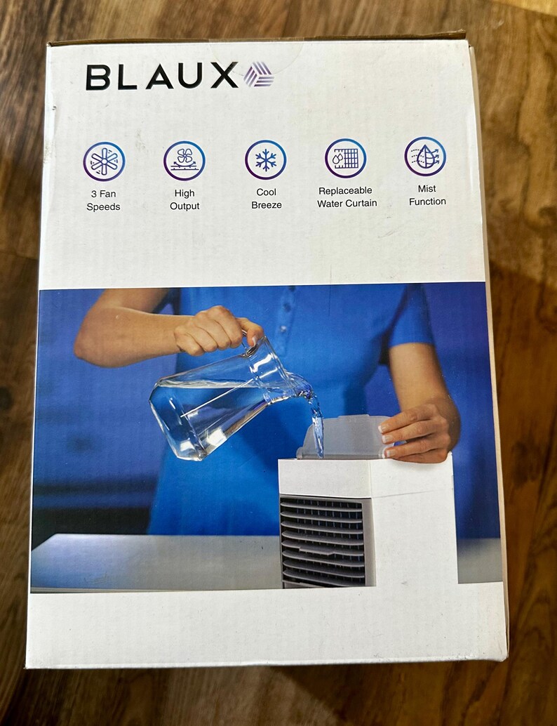 Puede incluir: Una caja de enfriador de aire port&aacute;til blanca y negra con la marca "BLAUX" en la parte superior. La caja presenta iconos que indican 3 velocidades de ventilador, alto rendimiento, brisa fresca, cortina de agua reemplazable y funci&oacute;n de niebla. Una persona vierte agua en el enfriador.