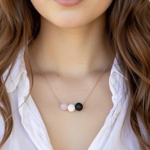 May include: A gold-colored necklace featuring three round beads: a pink, a white, and a black bead. The necklace is worn against a white collared shirt.
