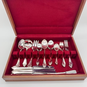 May include: A wooden storage chest with a deep red velvet interior containing a set of silver-toned flatware. The collection includes various spoons, forks, and knives with ornate handles, arranged in dedicated slots within the velvet-lined box.