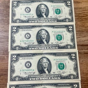 Pode incluir: São exibidas cinco notas de dois dólares americanos, cada uma com um retrato de Thomas Jefferson. As notas têm um selo verde e números de série, com a denominação claramente visível. As notas estão dispostas sobre uma superfície de madeira.