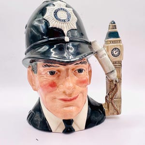 May include: A ceramic character jug shaped like a British police officer wearing a black helmet with a Metropolitan Police badge. The handle is designed as the Big Ben clock tower, featuring a clock face and a telephone receiver attached to the side.