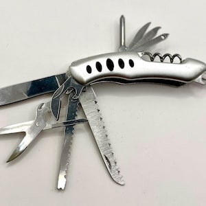 May include: A multi-tool with a silver handle and multiple fold-out tools, including a knife, scissors, saw, and corkscrew. The handle has five black oval cutouts. The tools are open and displayed against a white background.