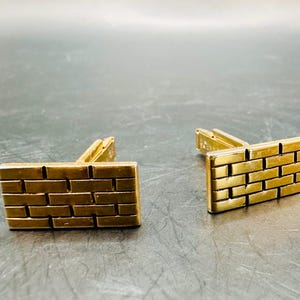 May include: A pair of gold-toned cufflinks with a brickwork design. Each cufflink is rectangular with a series of horizontal and vertical lines creating the brick pattern. The cufflinks are set against a dark background.