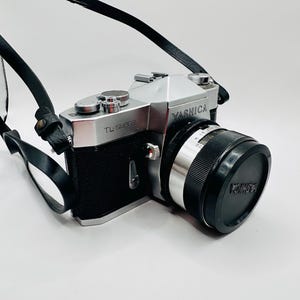 May include: A silver and black Yashica TL-Super 35mm film camera with a black leather strap. The camera body features the Yashica brand name and TL-Super model text. A black lens cap with the Yashica logo is attached to the front lens.