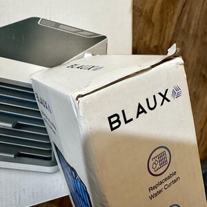 Puede incluir: Una caja de cart&oacute;n blanca con el nombre de la marca "BLAUX" en letras negras en negrita. La caja tambi&eacute;n presenta el texto "Replaceable Water Curtain" y un gr&aacute;fico de una cortina de agua. Otra caja con "AC ULTRA" es parcialmente visible.