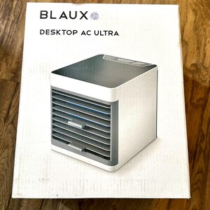 Puede incluir: Un enfriador de aire Desktop AC Ultra blanco y gris se muestra en su caja. La caja tiene el nombre de la marca BLAUX y el nombre del producto. El enfriador de aire tiene forma de cubo con ranuras horizontales en la parte delantera.