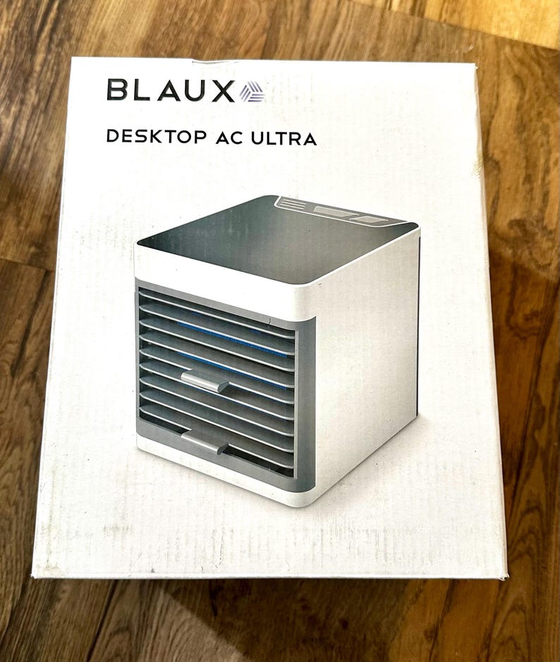 Puede incluir: Un enfriador de aire Desktop AC Ultra blanco y gris se muestra en su caja. La caja tiene el nombre de la marca BLAUX y el nombre del producto. El enfriador de aire tiene forma de cubo con ranuras horizontales en la parte delantera.