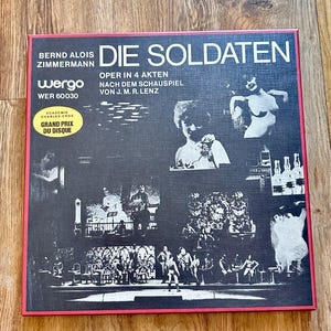 May include: A vintage vinyl record album cover with the title "Die Soldaten" in large white letters. The cover features black and white images of people and scenes from the opera, with text in German and French. The album is framed in red.