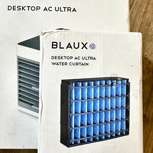 Puede incluir: Se muestran dos cajas blancas con el texto "BLAUX DESKTOP AC ULTRA". Una caja muestra un peque&ntilde;o aire acondicionado y la otra un filtro de cortina de agua. El filtro es negro con material filtrante azul.