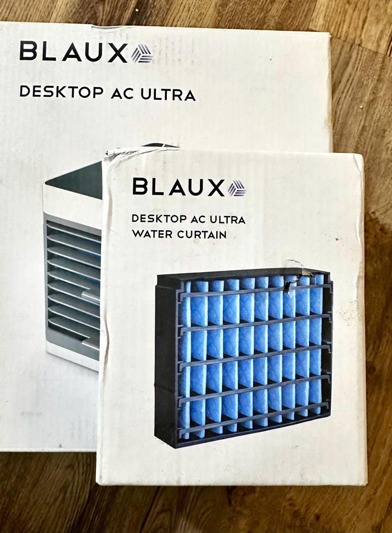 Puede incluir: Se muestran dos cajas blancas con el texto "BLAUX DESKTOP AC ULTRA". Una caja muestra un peque&ntilde;o aire acondicionado y la otra un filtro de cortina de agua. El filtro es negro con material filtrante azul.