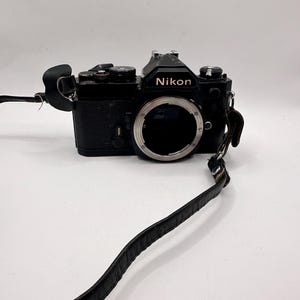 May include: A black Nikon film camera body featuring a central lens mount, a pentaprism housing with the Nikon logo in gold lettering, various top-mounted dials, and a black leather camera strap attached to the side lugs.